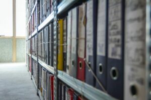 Binders and folders with archive documents are lined up along shelves in a storage room.