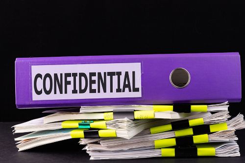 bigstock-Text-Word-Confidential-Is-Wri-370654939 A purple binder labeled "CONFIDENTIAL" rests on top of several stacks of paper documents clipped together with yellow and black binder clips, all set against a black background.